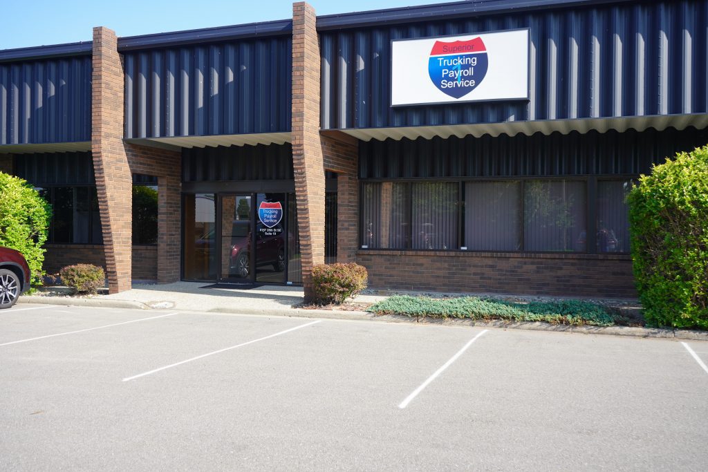 This image shows the exterior of a brick-and-black building with a sign for "Superior Trucking Payroll Service" above the entrance. The parking lot in front of the building is empty, and there are shrubs and greenery along the sidewalk near the windows and doors.