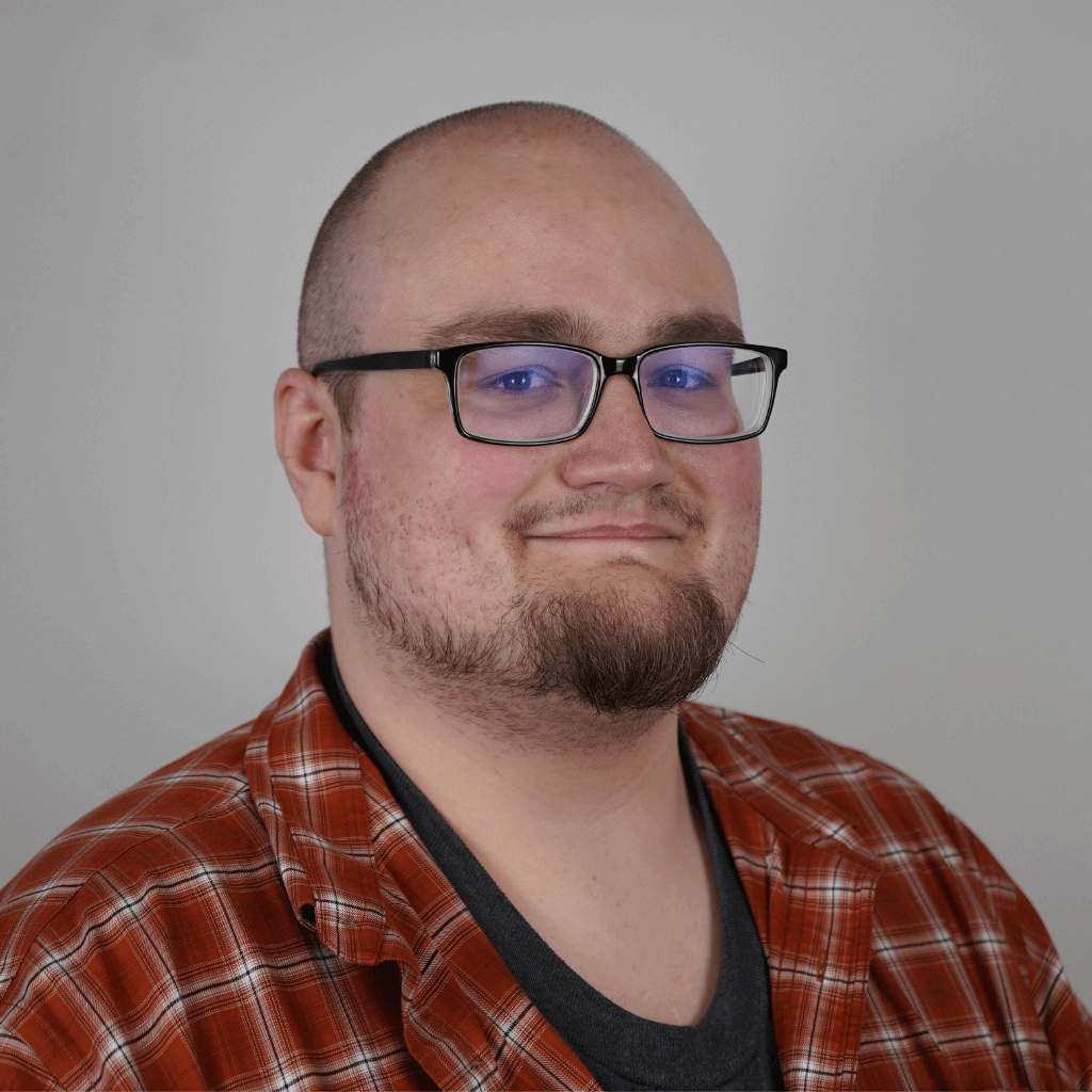 The image features Caleb with a shaved head, glasses, and a short beard. He is wearing a red and white plaid shirt over a dark gray T-shirt and is smiling subtly at the camera against a plain background.