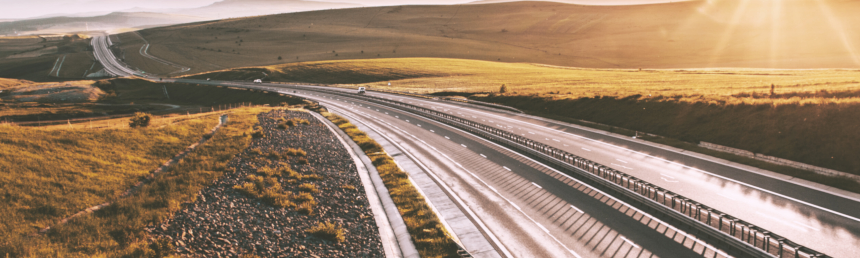 The image shows a long, empty highway stretching through a hilly, golden landscape at sunset. The road winds gently through the rolling terrain, illuminated by warm sunlight, creating a peaceful and scenic atmosphere.