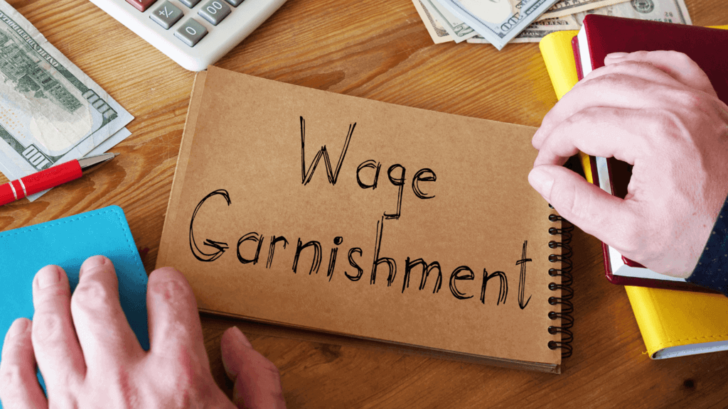 Notebook labeled “Wage Garnishment” on a desk surrounded by cash, calculator, and office supplies
