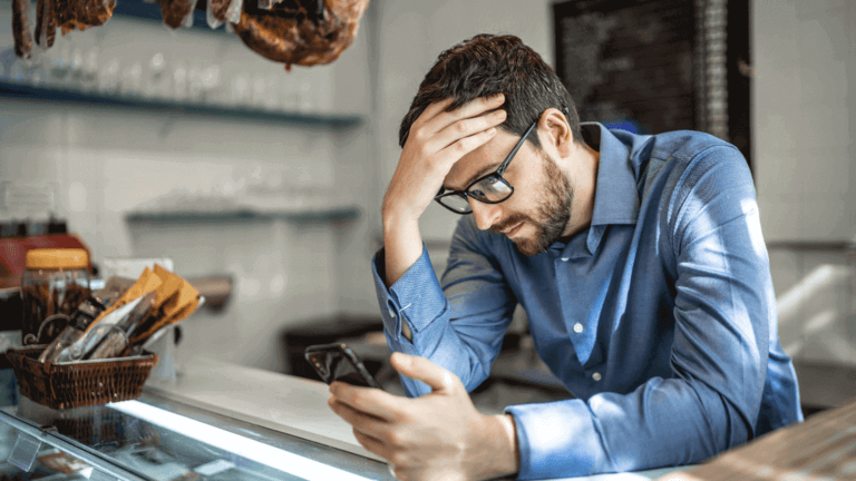 Man in a blue shirt looking stressed, holding his head with one hand and a phone with the other while standing behind a counter.